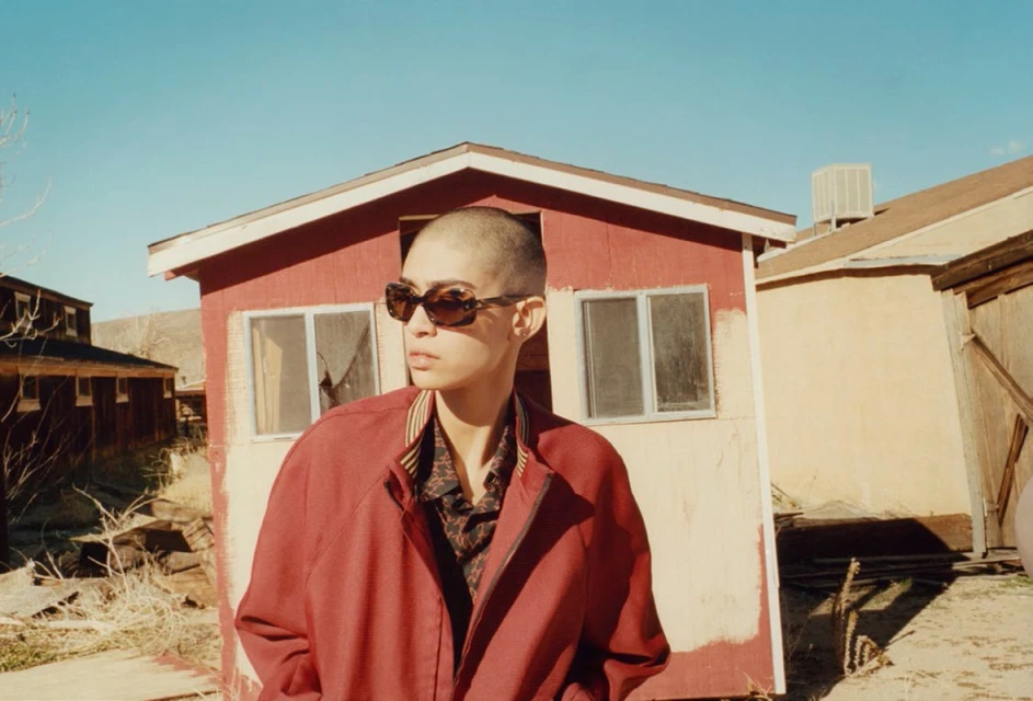 A young adult in sunglasses stands in front of a weathered red building with a tan roof against a blue sky in a desert landscape.