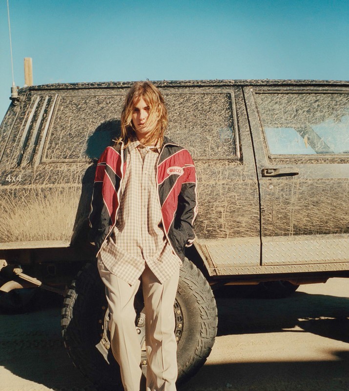 Adult male with long blond hair poses in casual clothing leaning against a dirty four-wheel drive car in a desolate outdoor setting.