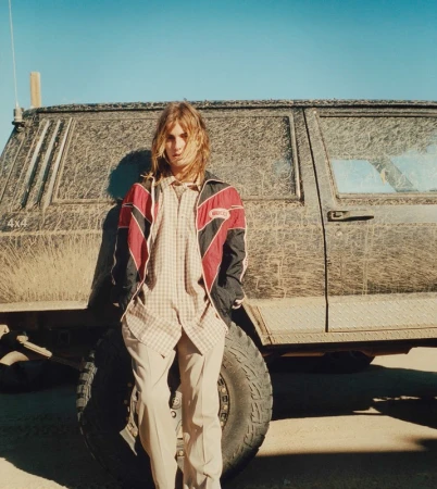 Adult male with long blond hair poses in casual clothing leaning against a dirty four-wheel drive car in a desolate outdoor setting.