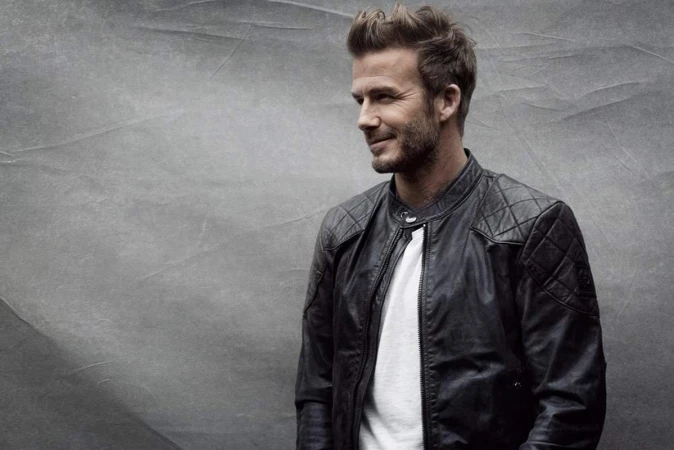 David Beckham wearing a black quilted leather jacket over a white t-shirt against a textured grey background