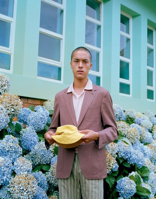 An adult male stands outdoors near hydrangea bushes, wearing a mauve suit and striped pants, holding a yellow hat, with a mint green building.