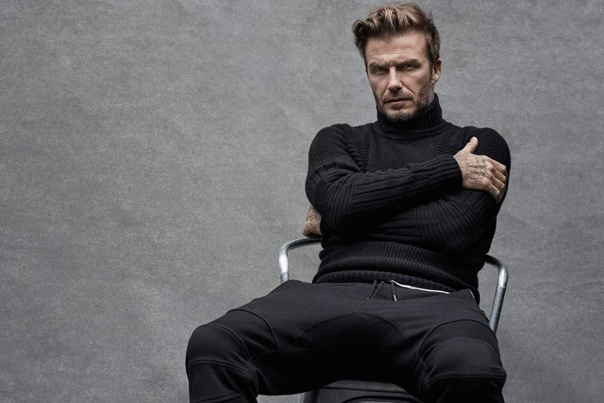 David Beckham in a black turtleneck and sweatpants sits on a metal chair against a grey backdrop in a medium shot, arms crossed.