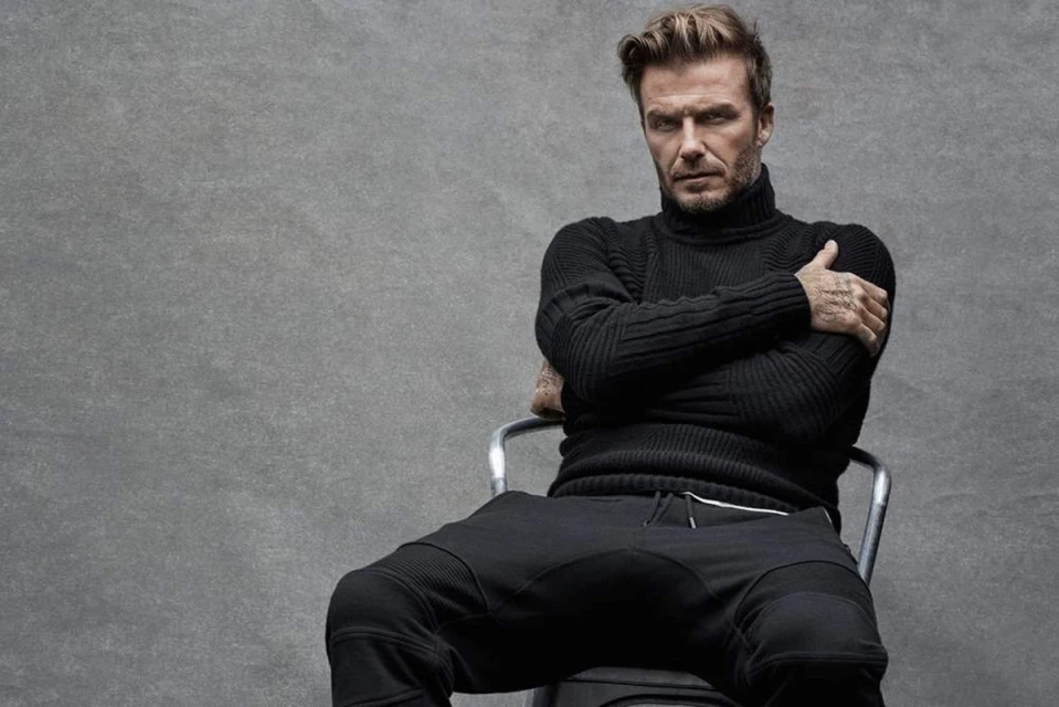 David Beckham in a black turtleneck and sweatpants sits on a metal chair against a grey backdrop in a medium shot, arms crossed. 