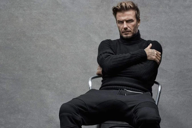 David Beckham in a black turtleneck and sweatpants sits on a metal chair against a grey backdrop in a medium shot, arms crossed. 
