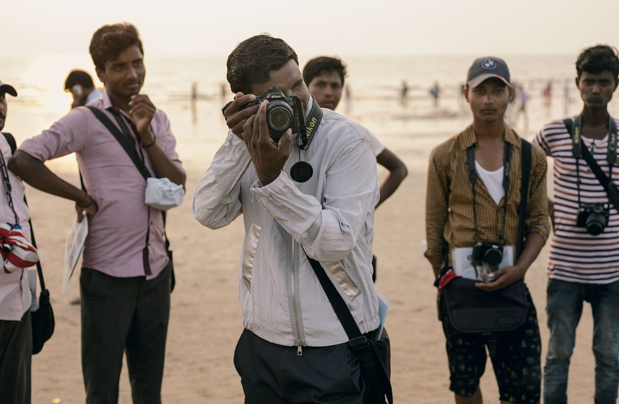 Group of photographers on a beach with cameras, sandy foreground, and ocean in the distance, wearing casual clothes in daylight.