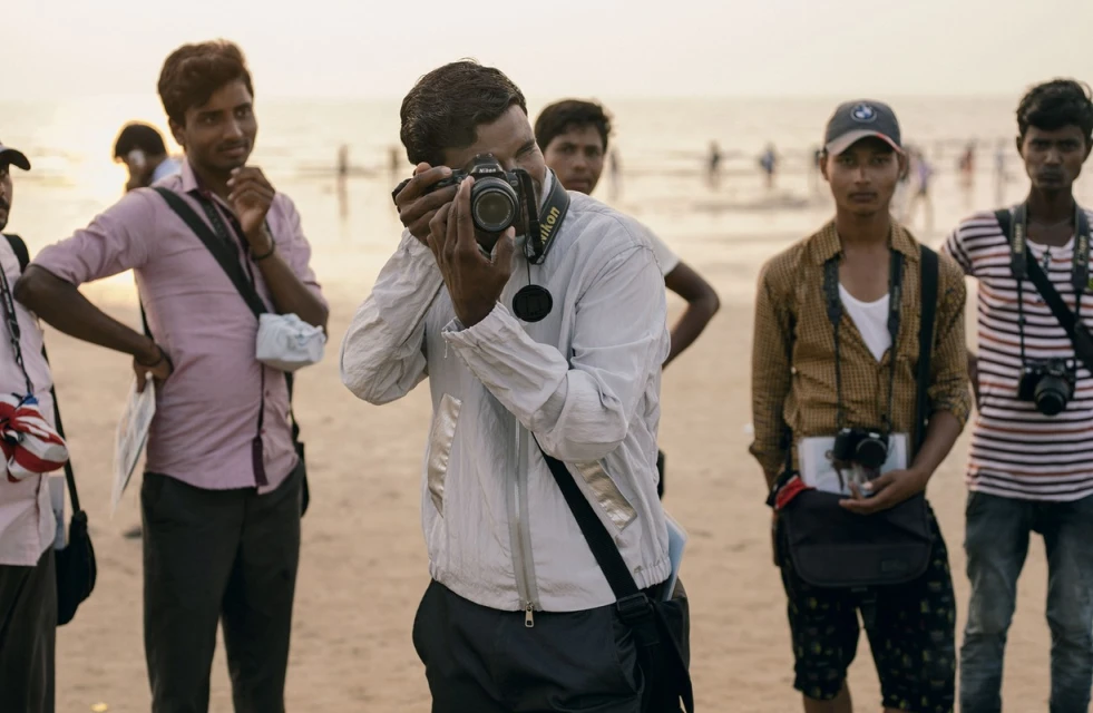 Group of photographers on a beach with cameras, sandy foreground, and ocean in the distance, wearing casual clothes in daylight.