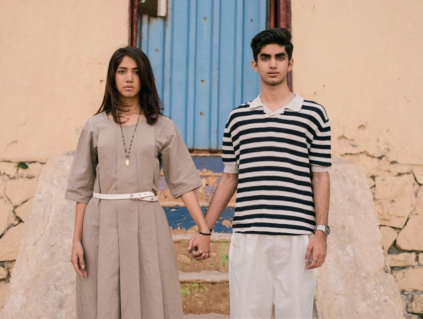 Two young adults, a male and a female, holding hands and standing in front of a building with a blue door, captured in a portrait shot.
