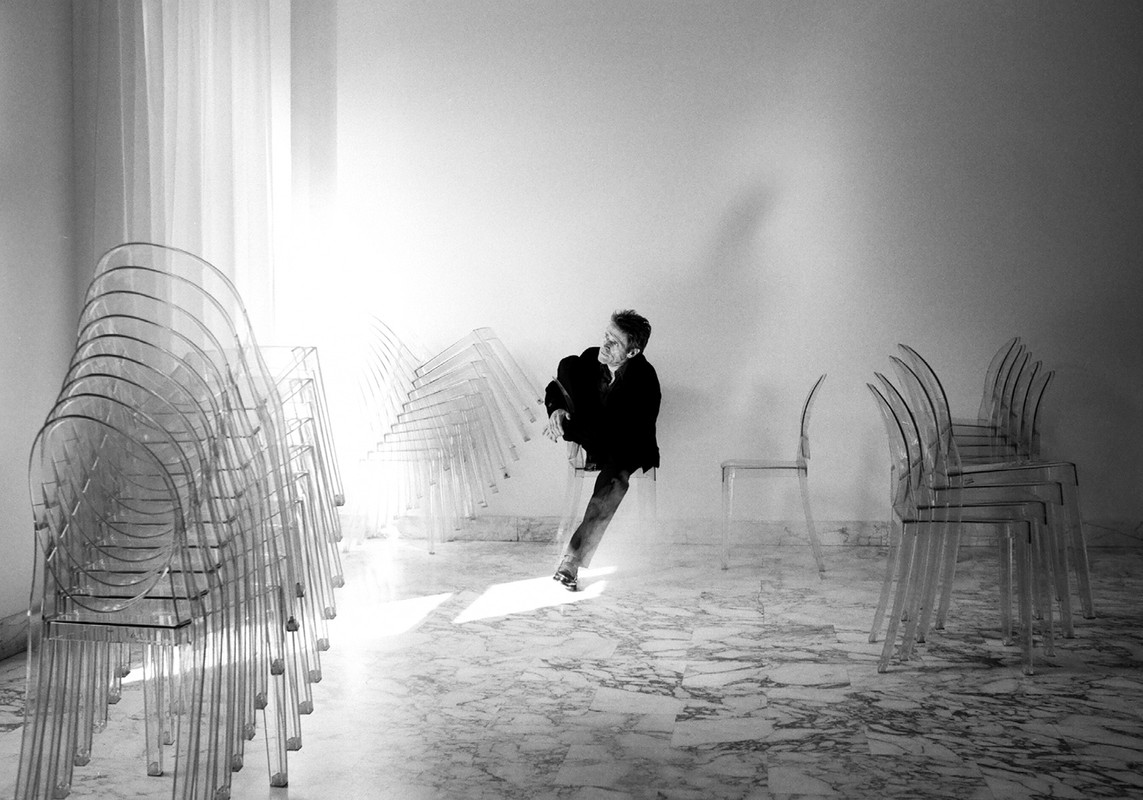 Monochrome shot of a man walking in a room with transparent chairs stacked on either side and a marble floor, capturing a sense of motion.