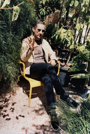 A man in sunglasses sits on a yellow chair amidst lush greenery, plants, and dappled light. He is wearing a tan jacket and dark pants.