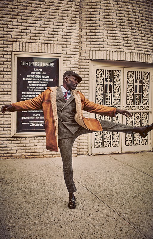 Man in tweed suit and tan coat strikes a pose with leg extended outdoors against a brick building in a full shot.