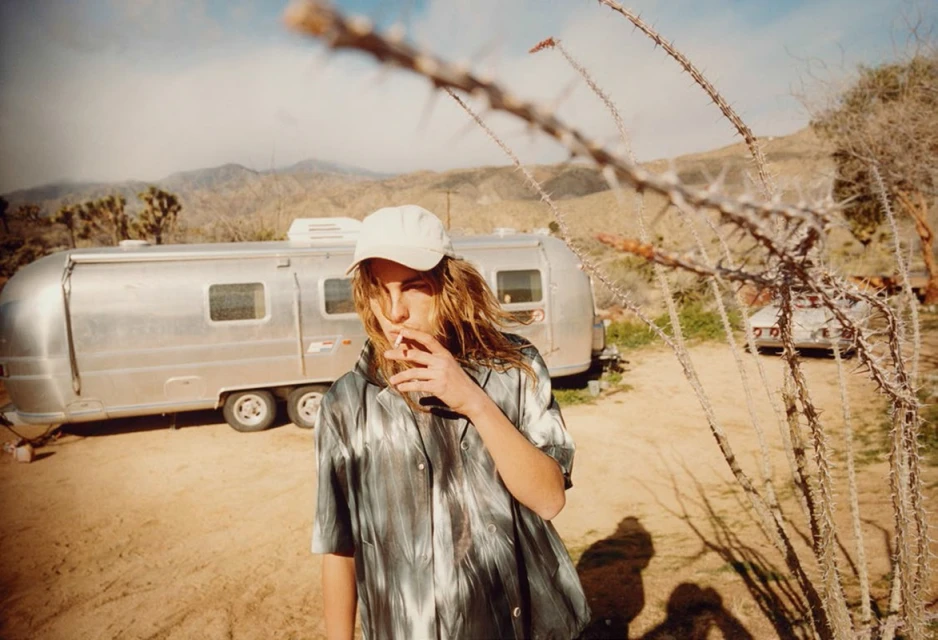 Adult wearing baseball cap smoking a cigarette in desert with travel trailer, arid landscape, and spiky desert plants in foreground.