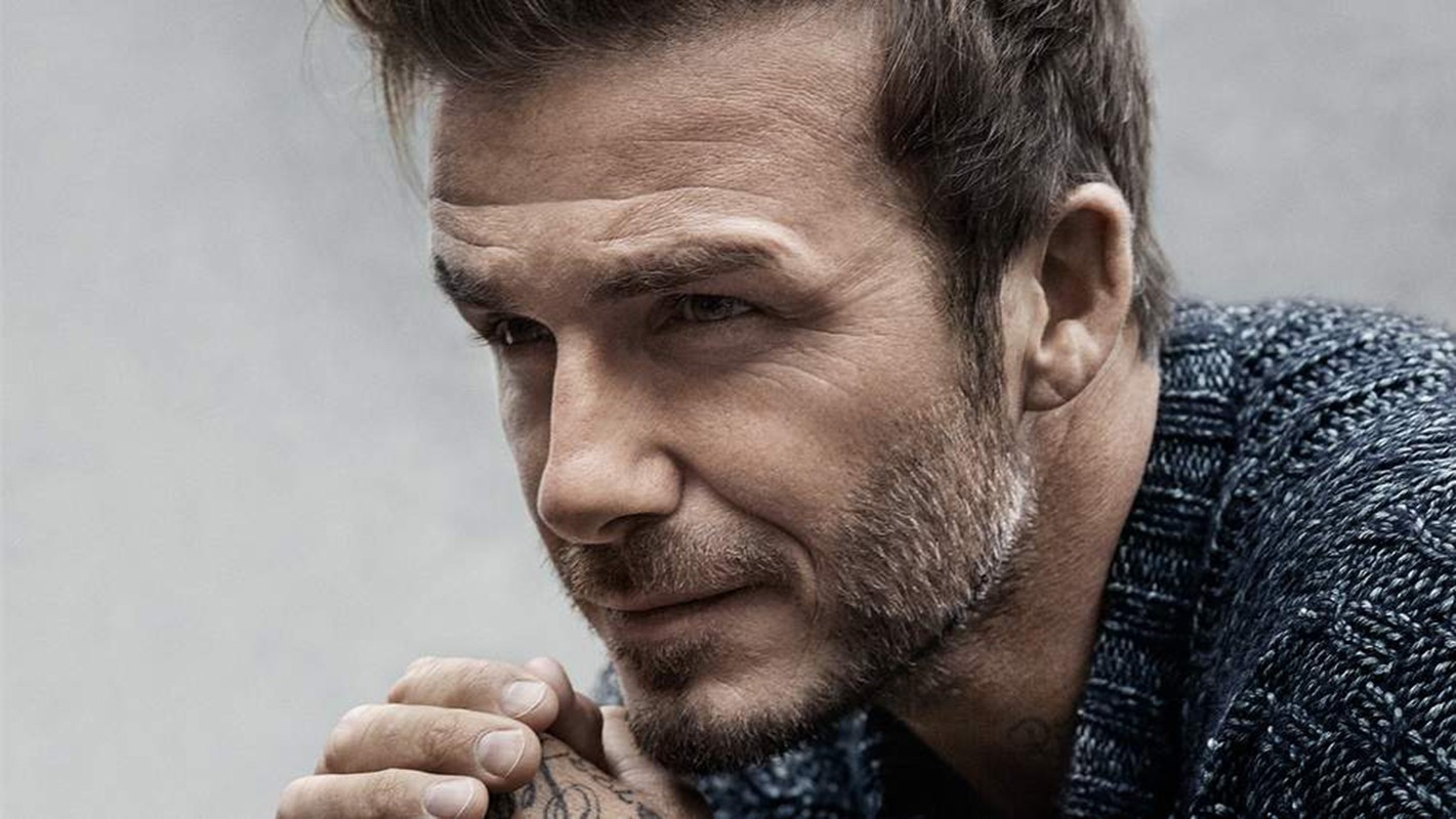 Close-up studio portrait of adult male with short dark hair, stubble beard, wearing dark knit sweater, hands clasped near chin, looking off-camera.