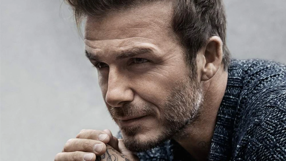 Close-up studio portrait of adult male with short dark hair, stubble beard, wearing dark knit sweater, hands clasped near chin, looking off-camera.