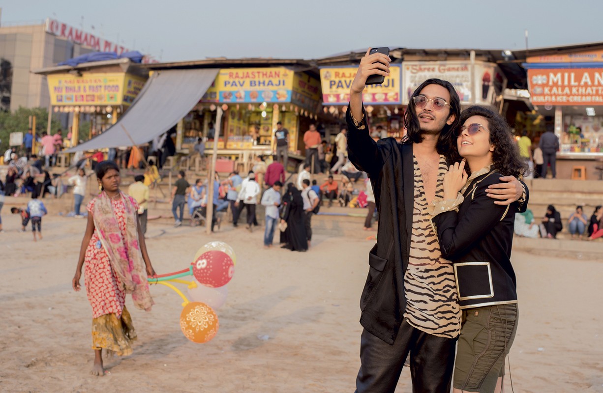 Couple poses for a selfie on a sandy beach with food stalls and people in the background during the day.