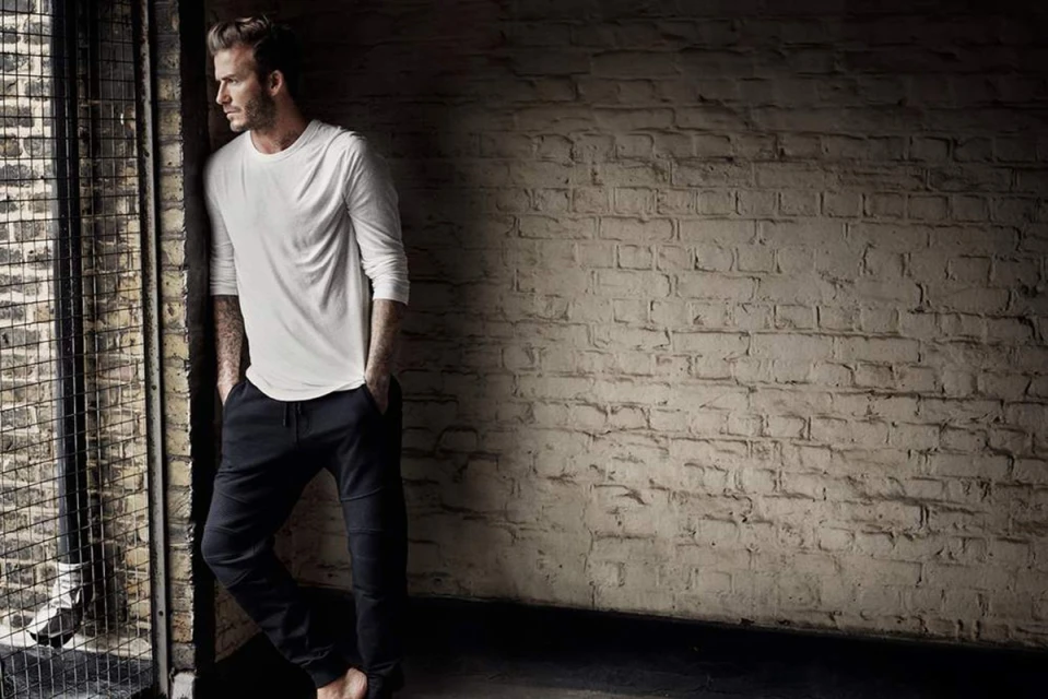 Adult man in white shirt and dark pants stands near a window with a metal grate in a dark room with a distressed brick wall.