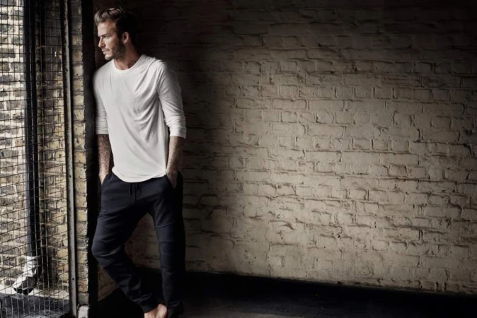 Adult man in white shirt and dark pants stands near a window with a metal grate in a dark room with a distressed brick wall.