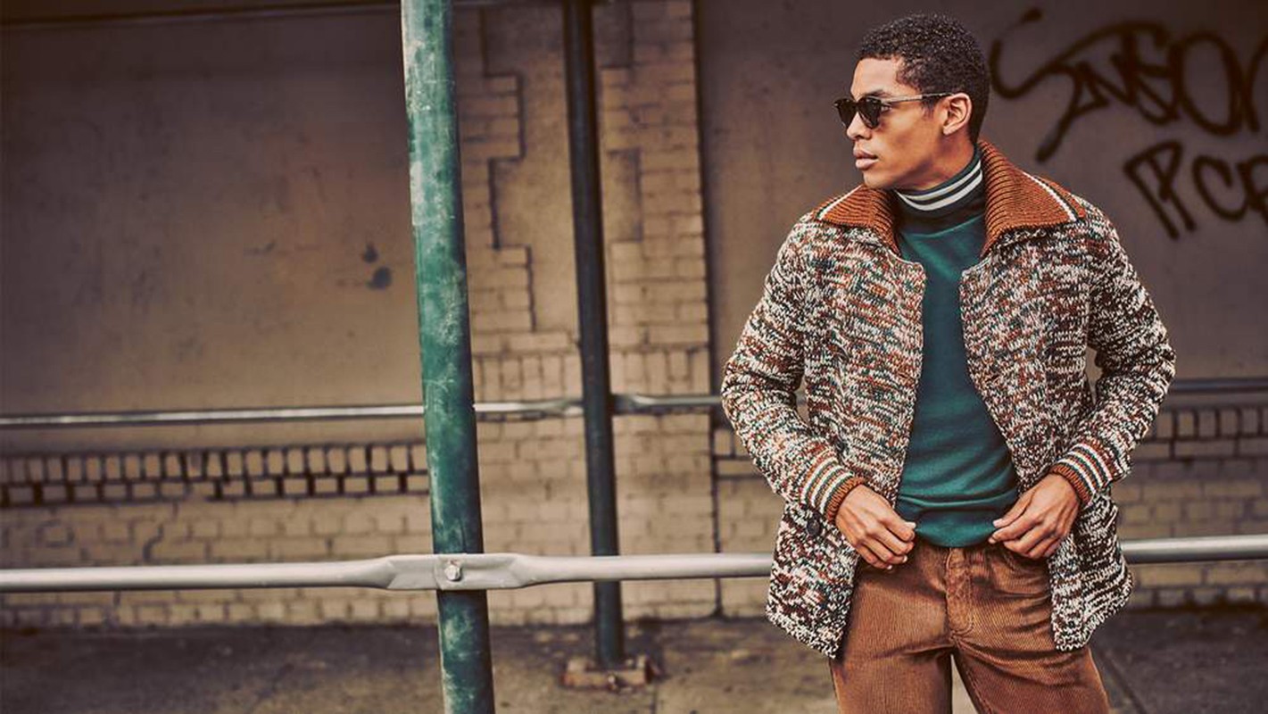 A man in sunglasses poses outdoors wearing a patterned cardigan, turtleneck, and corduroy pants next to metal bars, with graffiti in background.