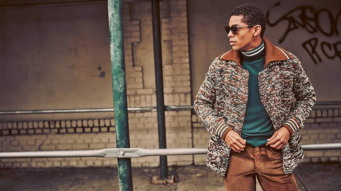 A man in sunglasses poses outdoors wearing a patterned cardigan, turtleneck, and corduroy pants next to metal bars, with graffiti in background.
