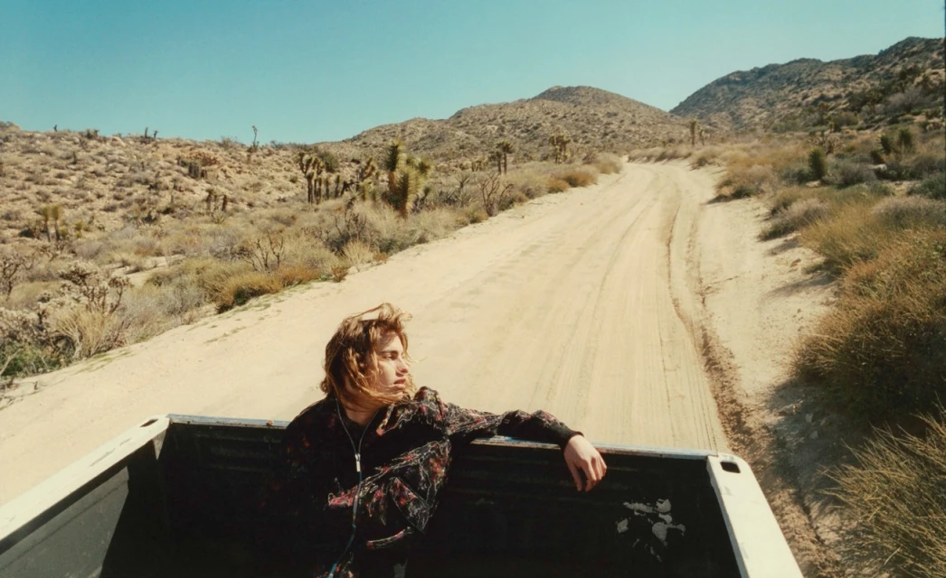 Young adult male in truck bed driving on a desert road with Joshua trees in the background on a sunny day with a clear blue sky.