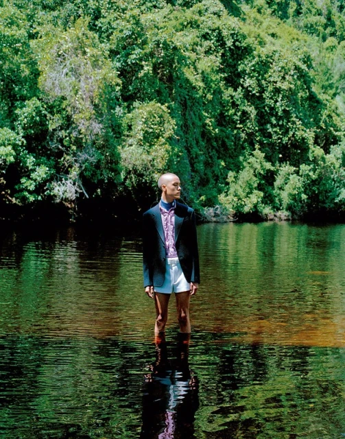 Bald adult man standing in shallow lake water, wearing a dark blazer, patterned shirt and white shorts with lush green forest backdrop.
