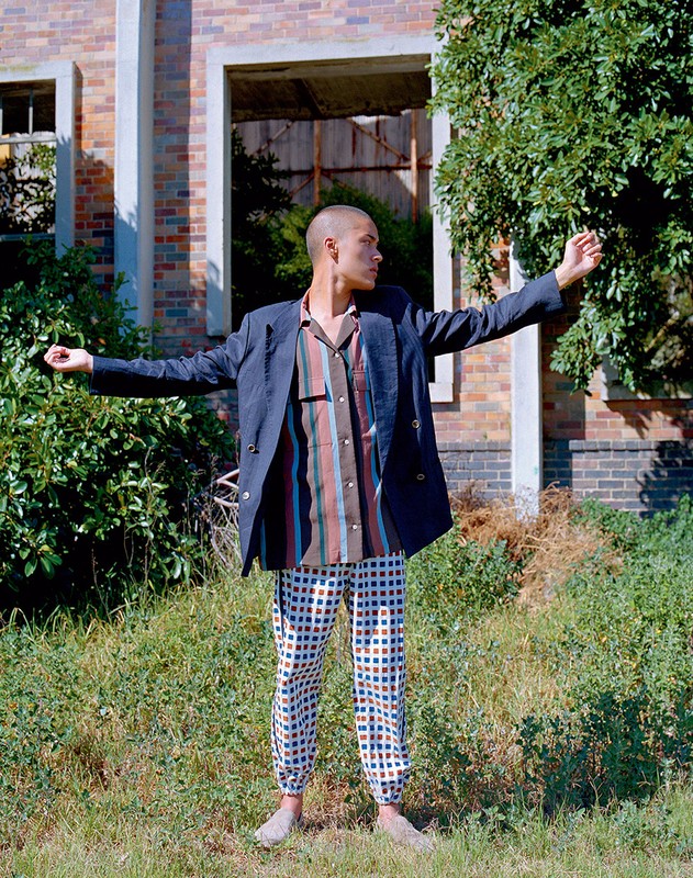 Adult male in a blue blazer and patterned pants stands in a grassy field in front of a brick building, posing with arms outstretched.