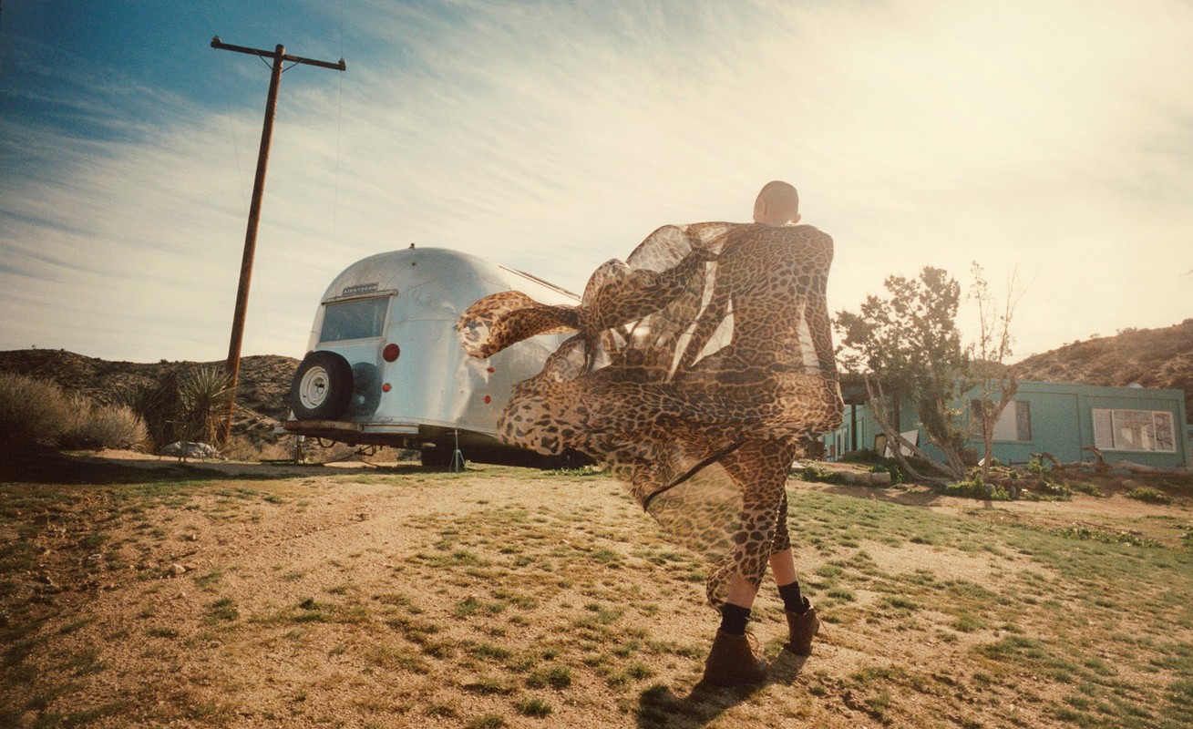 Woman in flowing leopard-print dress walks past Airstream trailer in desert landscape under a bright sky with scattered clouds.