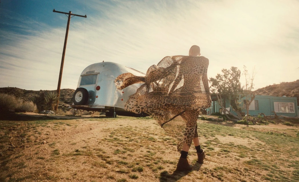 Woman in flowing leopard-print dress walks past Airstream trailer in desert landscape under a bright sky with scattered clouds.