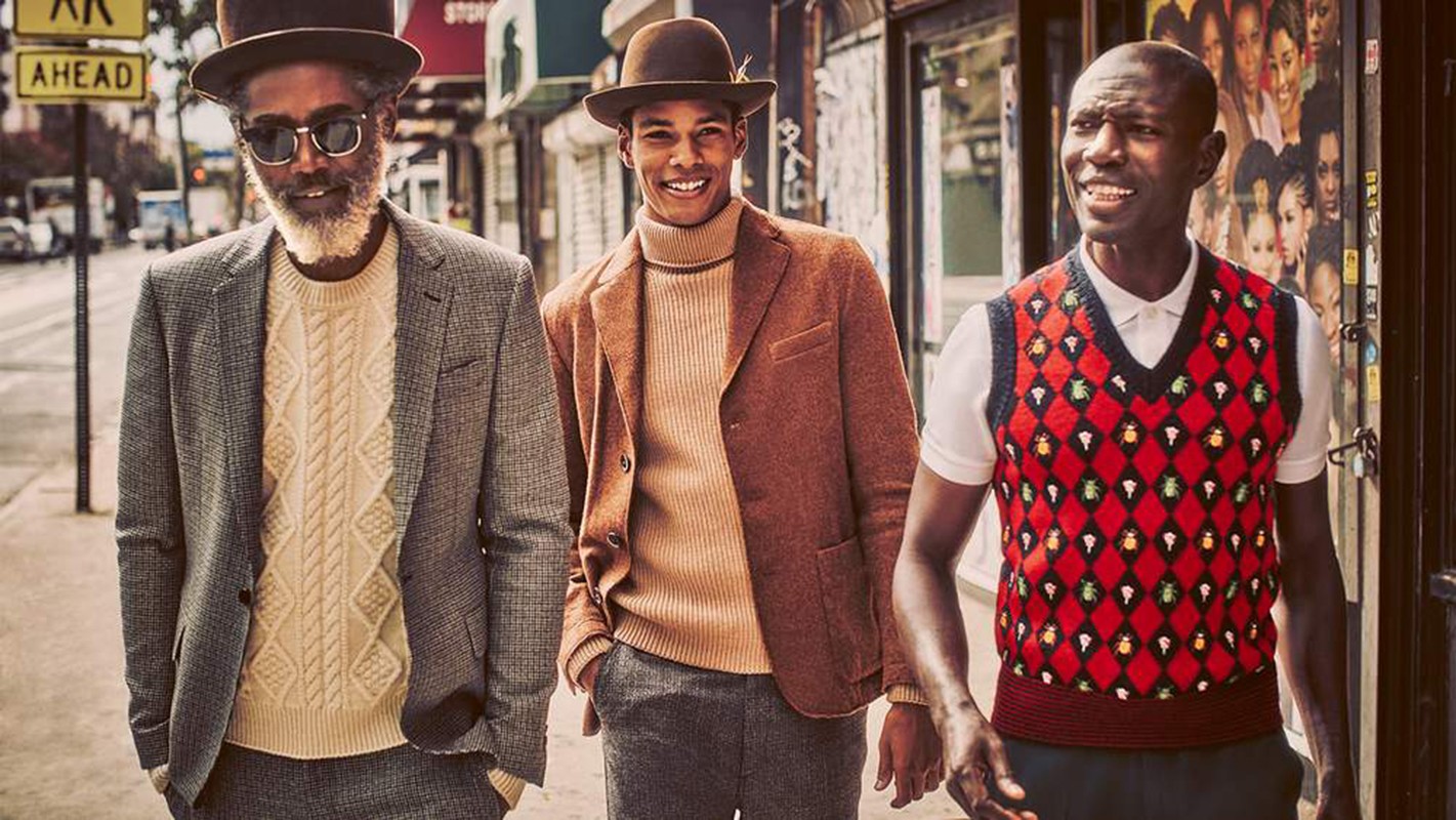 Three adult men walk on an urban sidewalk. One wears a grey blazer and hat, one wears a brown blazer and hat, one wears a sweater vest.