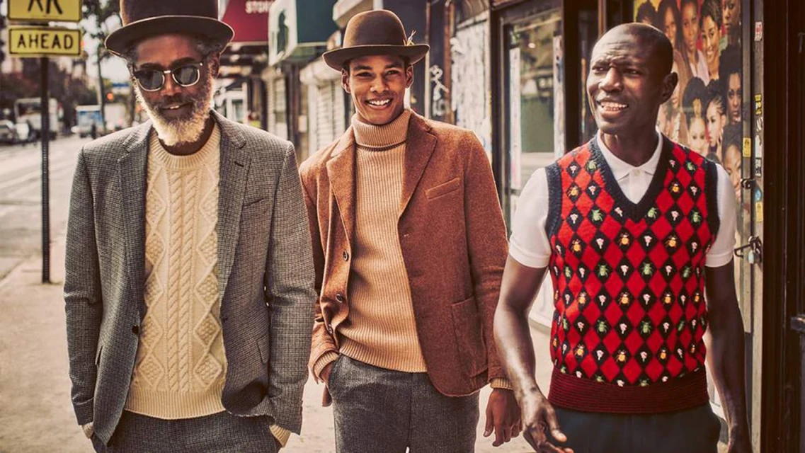 Three adult men walk on an urban sidewalk. One wears a grey blazer and hat, one wears a brown blazer and hat, one wears a sweater vest.