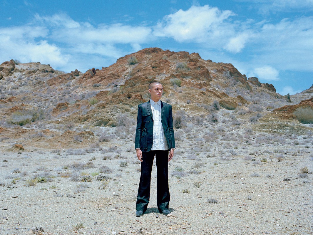 Adult male stands in a desert landscape wearing a jacket, pants, and dress shirt under a partly cloudy sky with low mountain range.