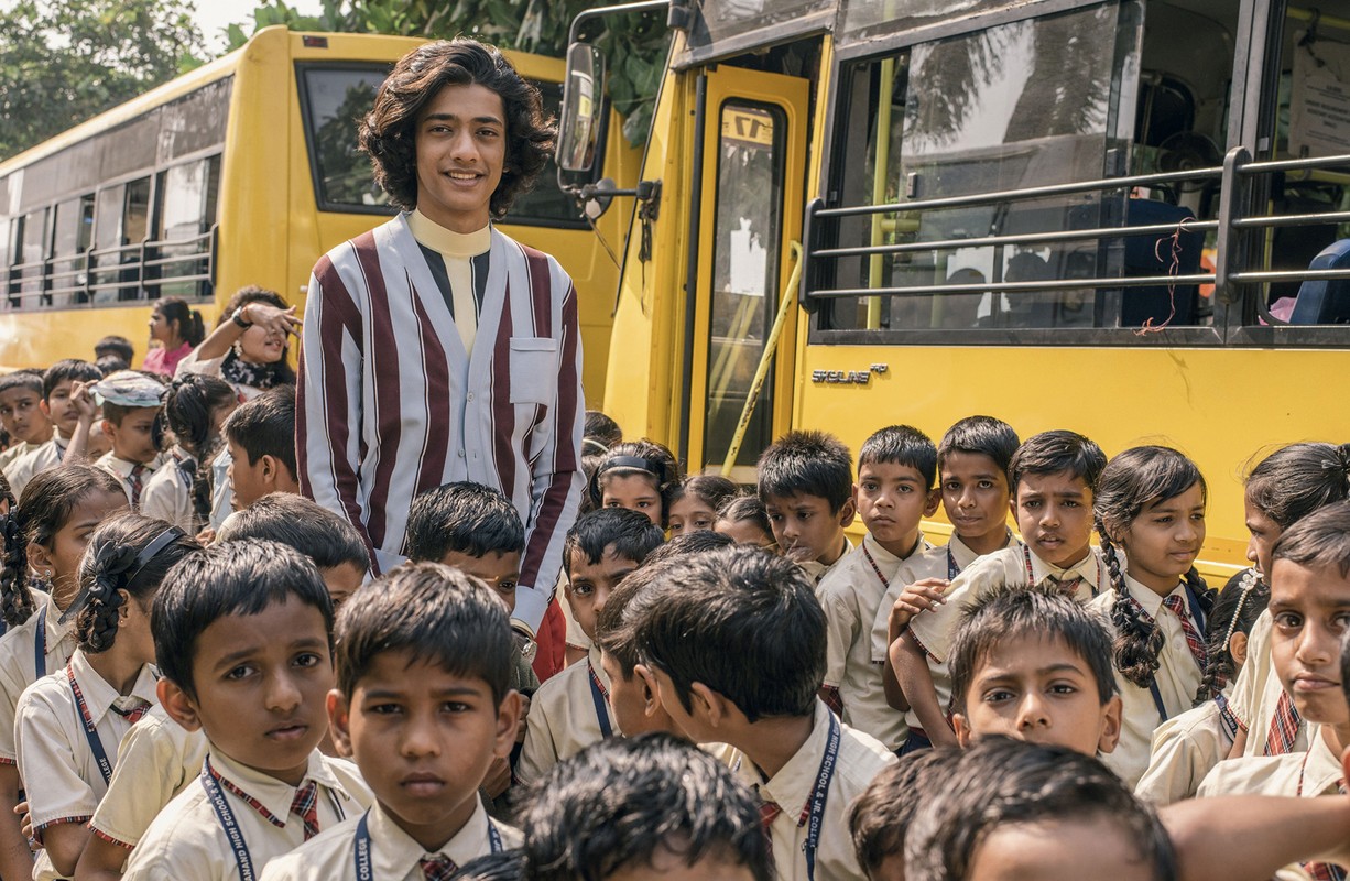 Young man in a striped shirt standing with a large group of children in school uniforms in front of a parked yellow school bus.