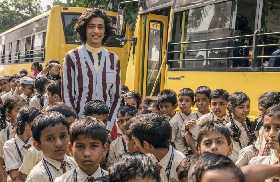 Young man in a striped shirt standing with a large group of children in school uniforms in front of a parked yellow school bus.