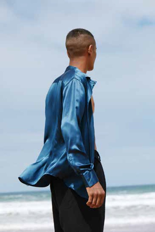 Man on a beach wearing blue satin shirt and black pants standing and looking at the ocean and blue sky in the daytime.
