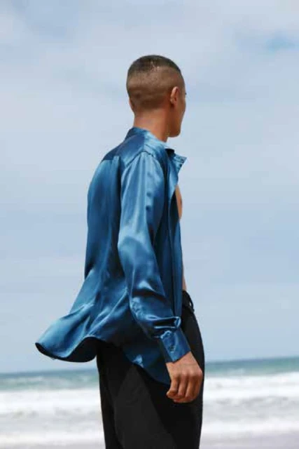 Man on a beach wearing blue satin shirt and black pants standing and looking at the ocean and blue sky in the daytime.