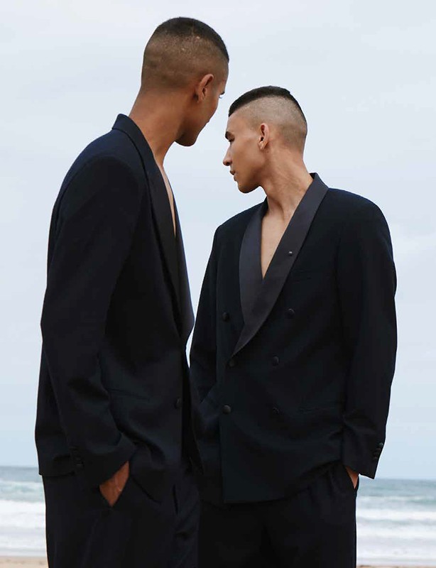 Two adult males in dark suits stand on a beach with the ocean and sky visible in the background. They are positioned close to each other.