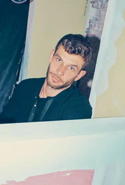 Portrait of a light-skinned adult male in a dark shirt, indoors, with a neutral expression and a textured wall in the background.