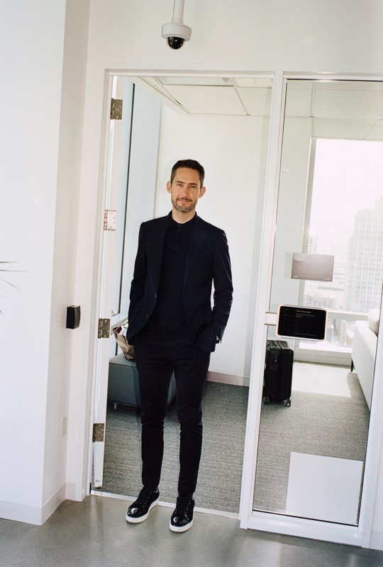 Indoor portrait of an adult male wearing a blazer and black clothing, standing in front of a doorway, with a security camera above.