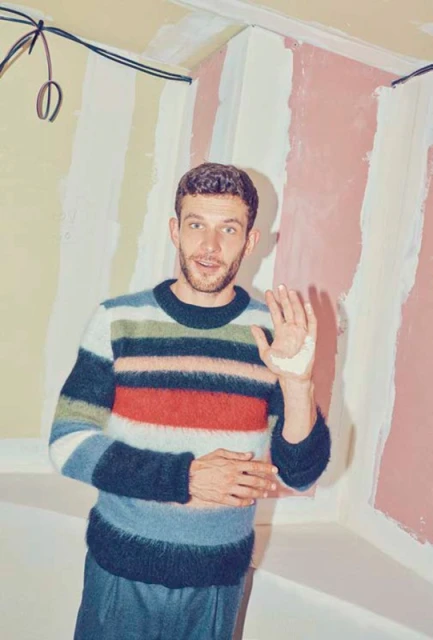 Man in striped sweater in interior with partially finished wall, smiling and holding up hand. Casual indoor scene. Fashion shot.
