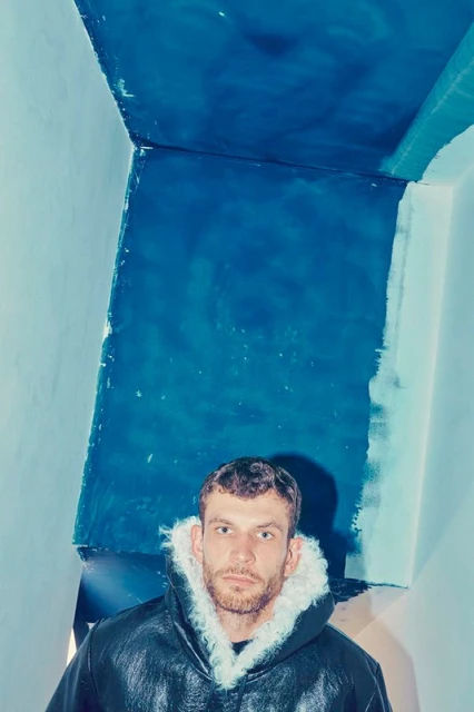 Portrait of a fair-skinned adult male with a beard, wearing a black fur-lined coat, standing indoors with painted blue walls.
