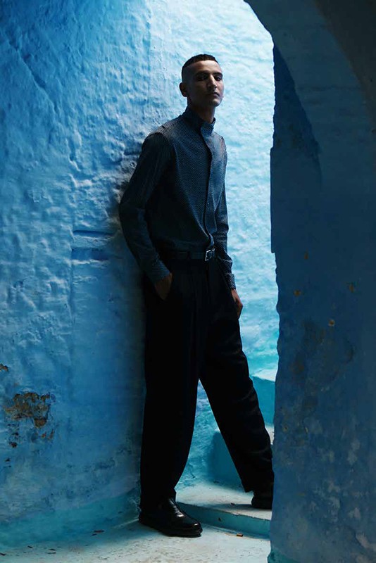 Man in a casual button-down and black pants standing in a blue stone archway with a neutral facial expression and his hand in his pocket.