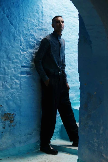 Man in a casual button-down and black pants standing in a blue stone archway with a neutral facial expression and his hand in his pocket.