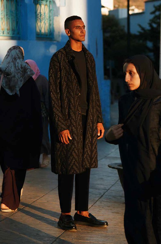 Man wearing a textured black patterned long coat and dark loafers standing outdoors with other people nearby.