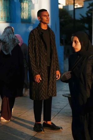 Man wearing a textured black patterned long coat and dark loafers standing outdoors with other people nearby.