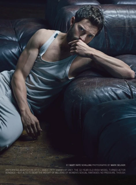 Jamie Dornan in studio portrait leaning against leather couch in gray pants and tank top looking at the camera with his hand near his mouth.