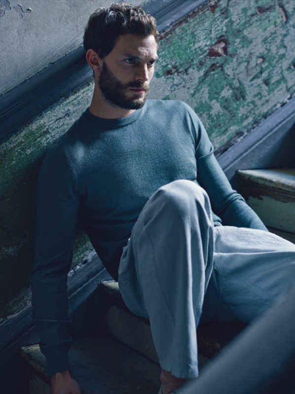Man with beard and blue sweater sitting on worn, weathered wooden stairs indoors in natural light. Fashion or celebrity portrait.