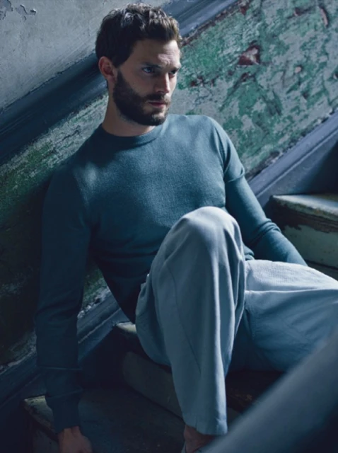 Man with beard and blue sweater sitting on worn, weathered wooden stairs indoors in natural light. Fashion or celebrity portrait.