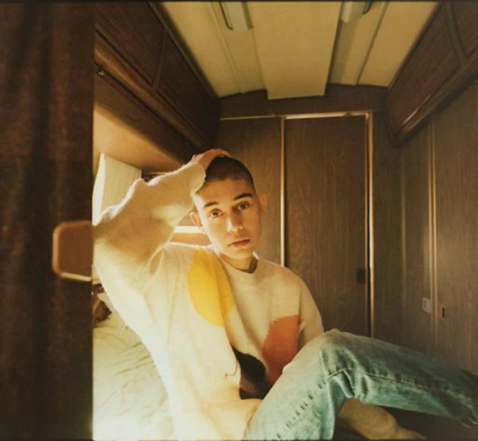 Young adult sits in a camper van, wearing a colorful sweater and jeans, with wood paneling and simple lighting in the background.