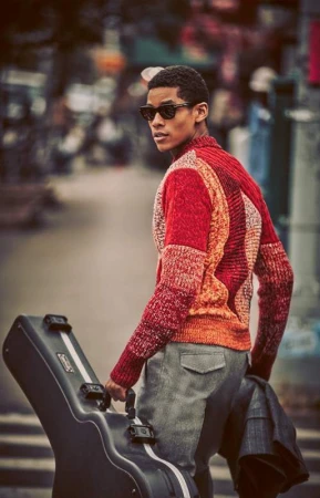 A stylish African-American man with sunglasses and a colorful sweater carries a guitar case in a street scene with blurred background.