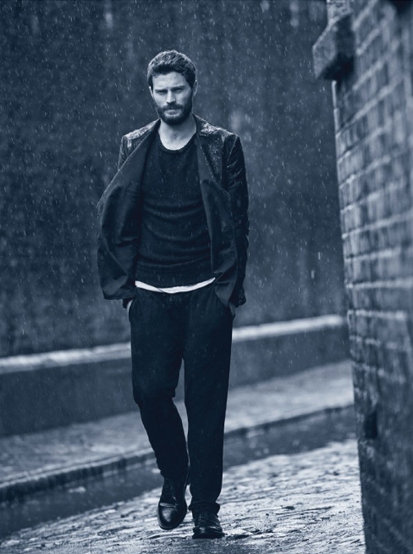 An adult male with a beard walks on a wet brick path in the rain, wearing a black suit and matching shirt and shoes, with brick walls.