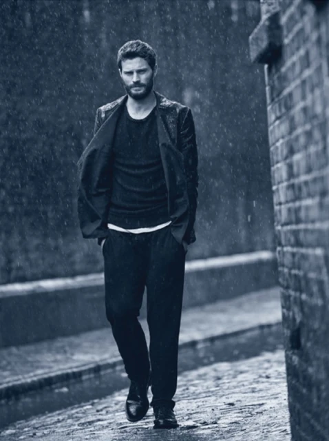An adult male with a beard walks on a wet brick path in the rain, wearing a black suit and matching shirt and shoes, with brick walls.